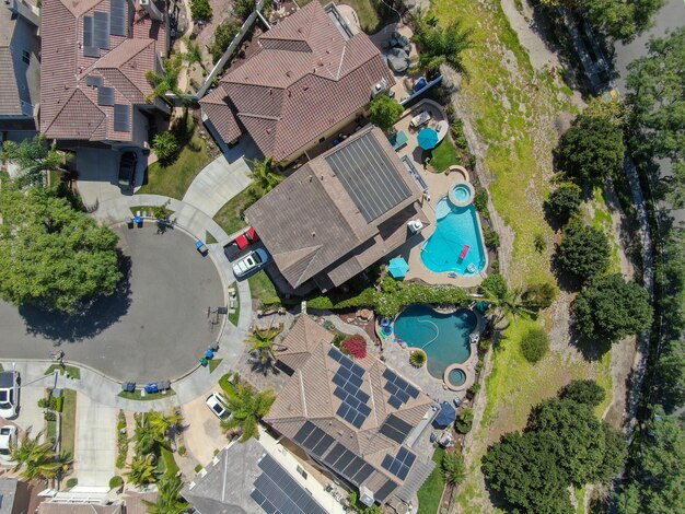 A drone shot of a residential neighborhood with solar panels installed on rooftops. A few houses have battery storage systems installed next to electrical panels. The image showcases the adoption of residential battery storage for grid stability.
