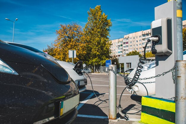 A charging station with multiple electric vehicles plugged in, with a backdrop of residential houses and a clear blue sky, indicating the integration of electric vehicles into daily life.